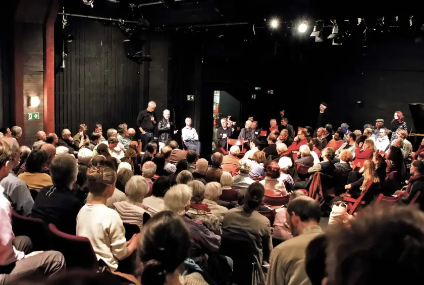 Une salle de théâtre remplie de spectateurs attentifs, avec des intervenants sur scène.