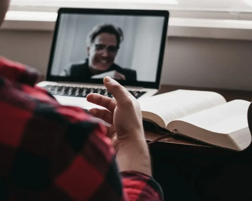 Une personne en chemise à carreaux discute en visioconférence sur un ordinateur portable, avec un livre ouvert à côté.