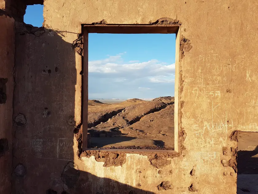 Vue à travers une fenêtre en ruine, montrant un paysage désertique sous un ciel bleu.