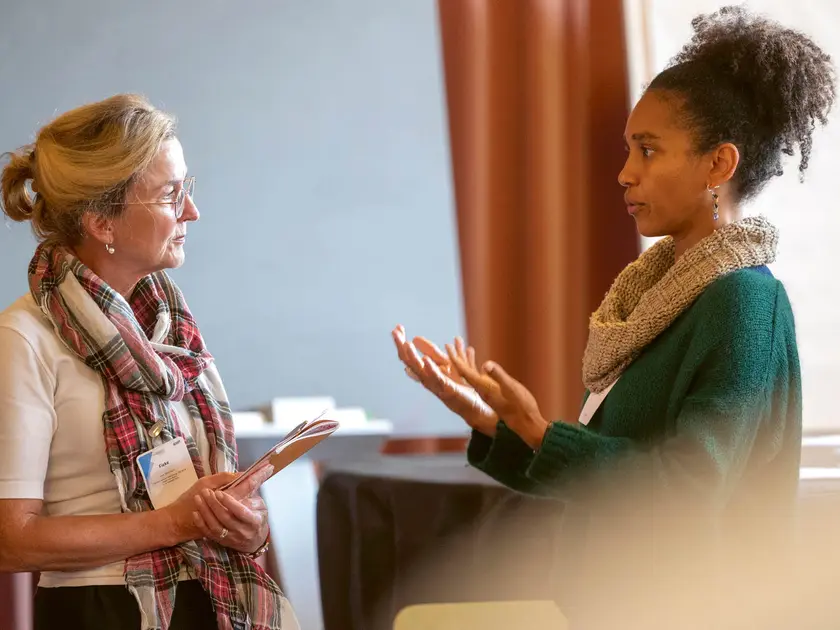 Deux femmes en discussion, l'une avec des lunettes et une écharpe, l'autre avec des cheveux frisés, dans un cadre intérieur.
