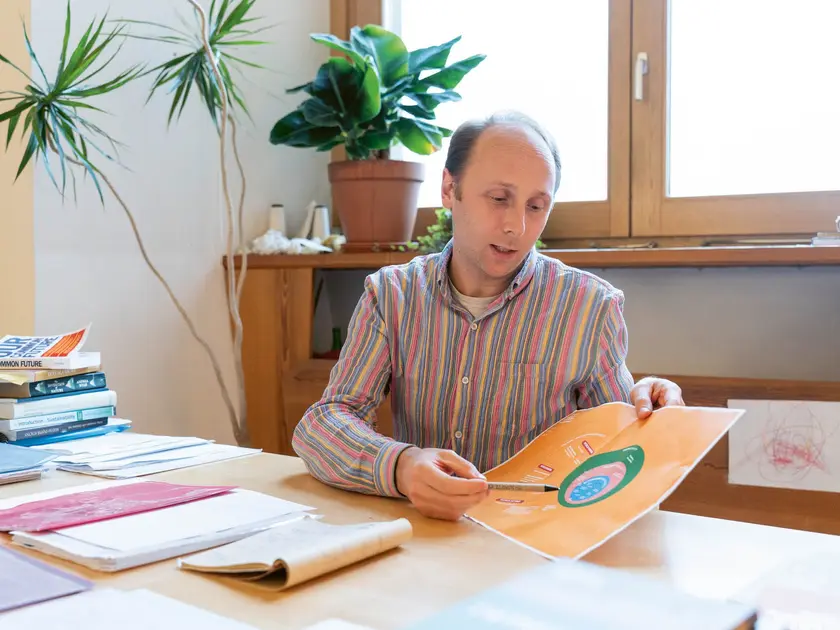 Un homme assis à une table, montrant un graphique coloré, entouré de livres et de plantes.
