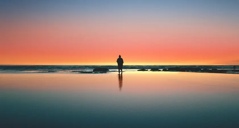 Silhouette d'une personne se tenant sur une plage au coucher du soleil, reflet dans l'eau.