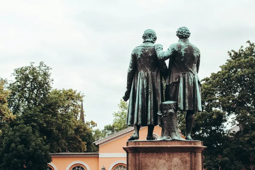 Deux statues en bronze représentant des hommes de dos, entourées d'arbres et d'un bâtiment en arrière-plan.
