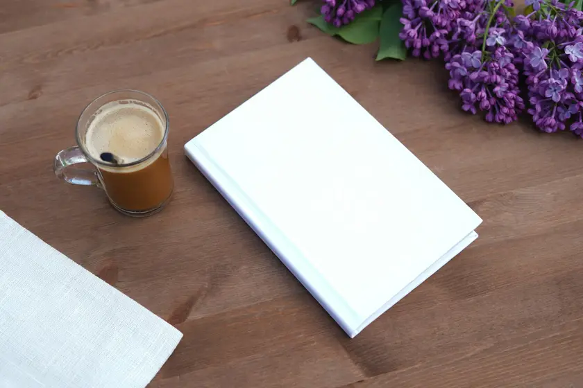 Un livre blanc posé sur une table en bois, accompagné d'une tasse de café et de fleurs lilas.