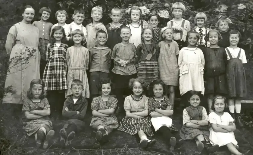Groupe d'une trentaine d'enfants posant en deux rangées avec une femme adulte, en extérieur devant un buisson, photographie noir et blanc ancienne.